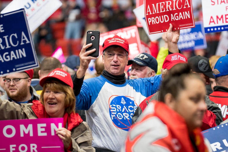 A man wearing a Space Force shirt and a MAGA hat holds his phone up to document the crowd at a Trump rally. The energetic crowd around him holds pro-Trump signs.